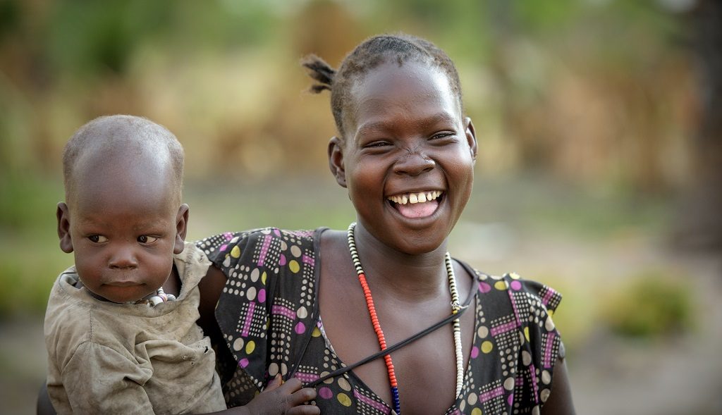 Mother and baby watch as other community members water their vegetable gardens early in a Saturday morning in Kuajok, Warrup State, South Sudan.